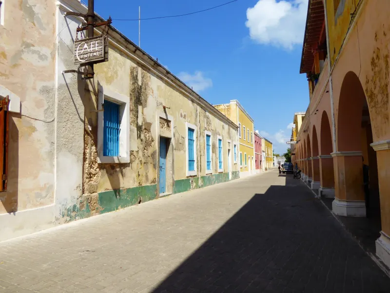 Weathered lime-washed facades with blue shutters along a paved street on the Island of Mozambique, an arcade opposite.
