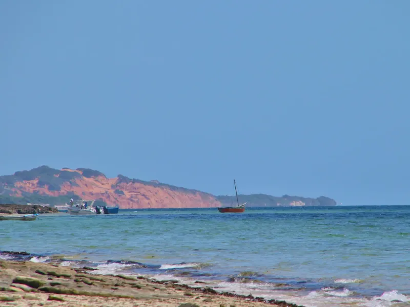 Red dune cliffs across the water at Vilankulo, with a dhow and small boats anchored in the shallows under a clear sky.