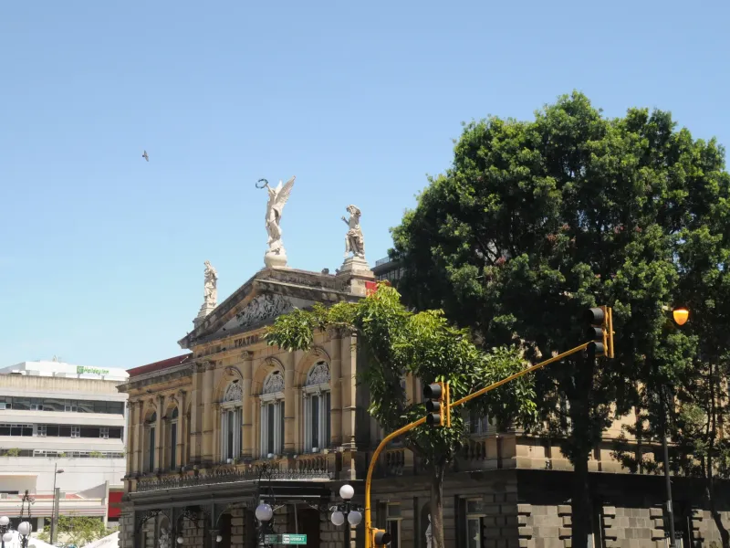 The columned stone facade and rooftop statues of the Teatro Nacional in San Jose, Costa Rica, seen past a street tree