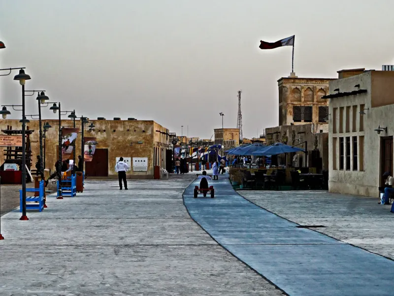 A paved waterfront promenade at Souq Al Wakrah lined with coral stone buildings and a Qatari flag