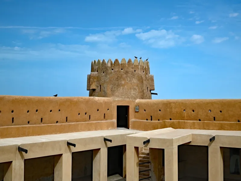 The restored mud brick fort at Al Zubarah, with a crenellated wall and corner tower against a clear sky