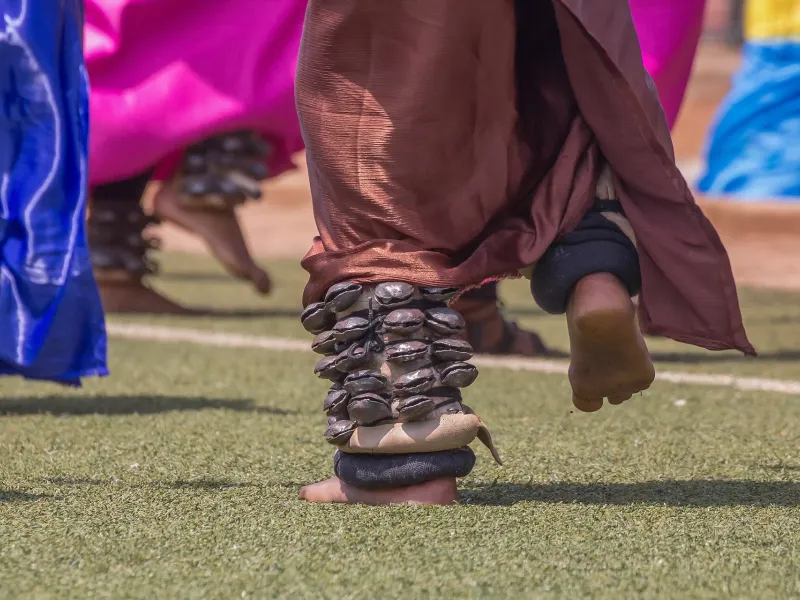 Close view of Intore dancers mid-step, seed-pod amayugi bells bound at their ankles over brightly colored wraps, on a pitch in Rwanda.