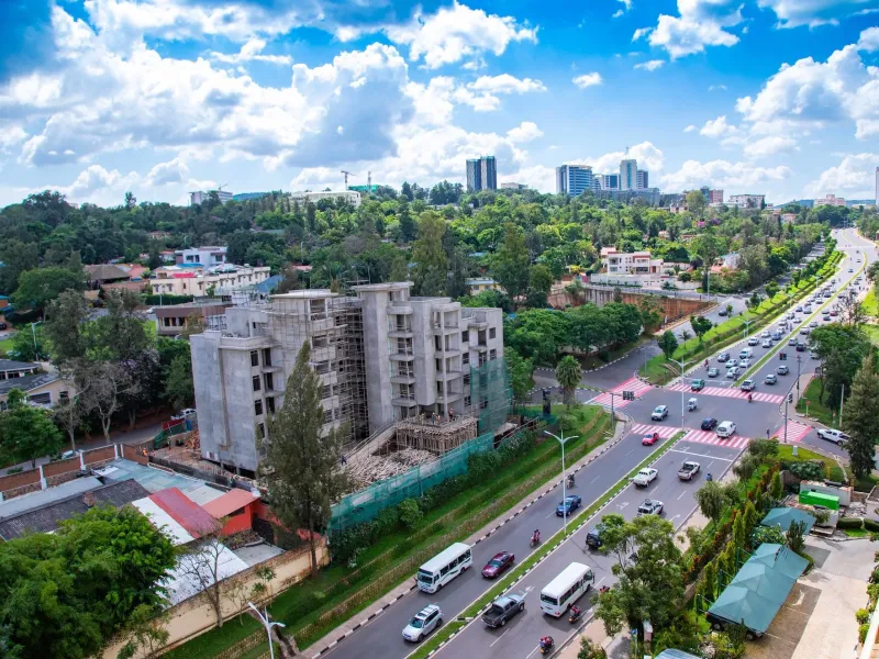 A divided boulevard in Kigali seen from above, with minibuses, motorcycle taxis and cars, a building under construction, and the downtown towers on the ridge behind.