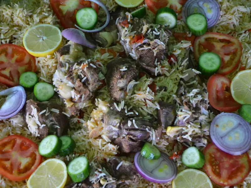 A shared platter of kabsa, spiced rice with meat, dressed with sliced tomato, cucumber, onion and lemon.