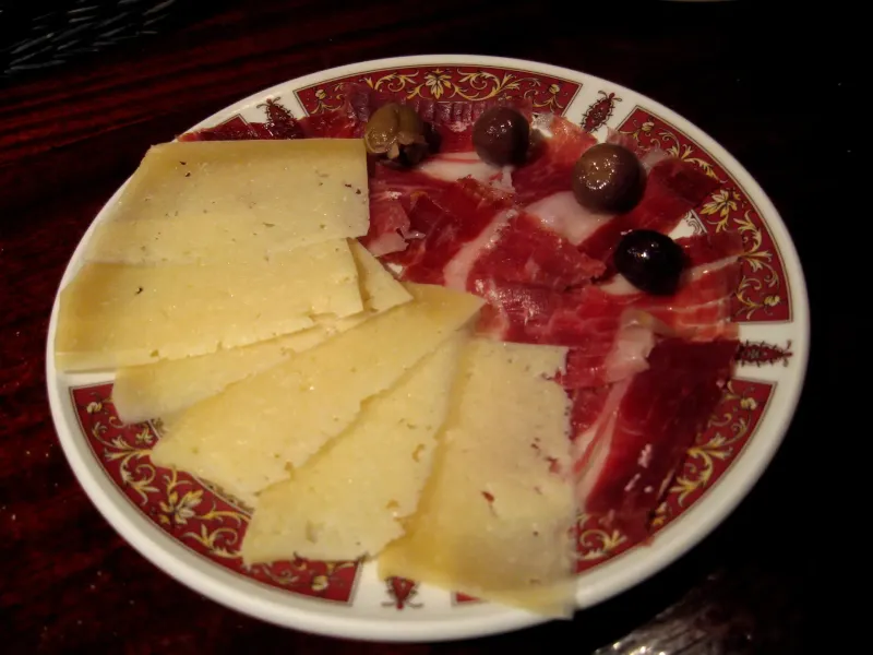 A plate of cured ham slices, wedges of hard cheese and dark olives served as a tapa in Seville, Spain.
