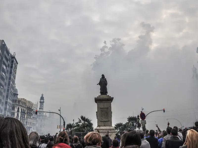 Firework smoke drifting over a statue and a packed crowd in the city square during a mascleta at Las Fallas in Valencia.