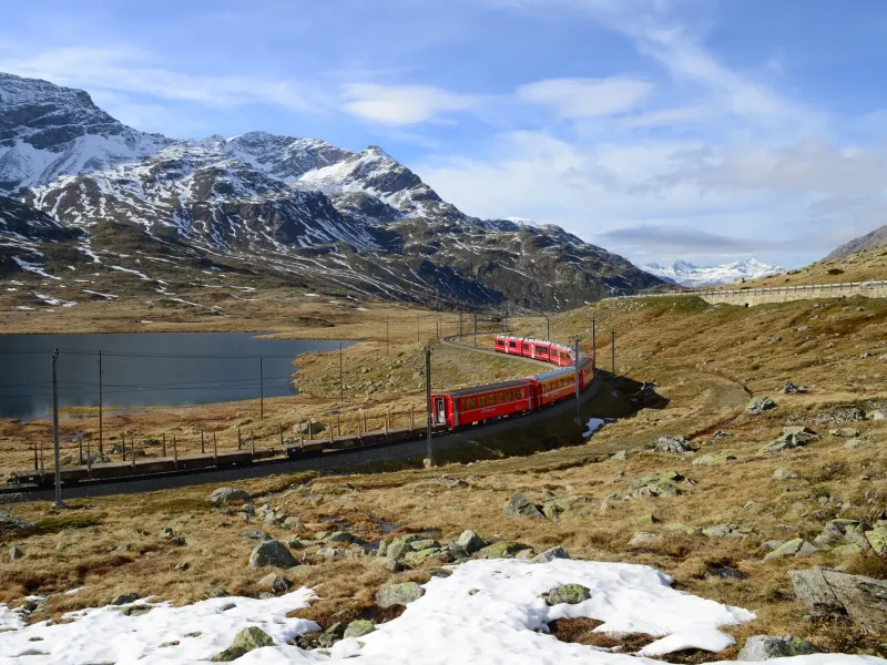 A red regional train running along the shore of Lago Bianco on the Bernina line, snow still lying on the high pasture.