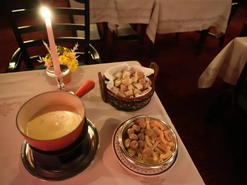 A caquelon of Swiss cheese fondue on its burner with bowls of cut bread and boiled potatoes beside it on a white cloth.