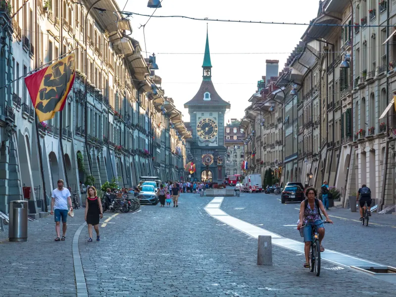 The Zytglogge clock tower at the end of a cobbled street in Bern's old city, with arcades and cantonal flags either side.