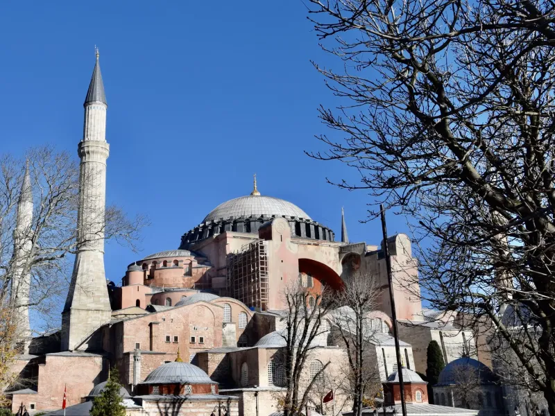 Hagia Sophia in Istanbul, its shallow dome and ochre buttressed walls above a stone minaret, through bare winter trees.