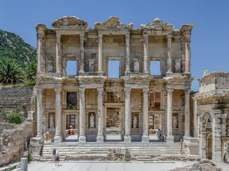 The two-story marble facade of the Library of Celsus at Ephesus, its columns and statue niches against a wooded hillside.