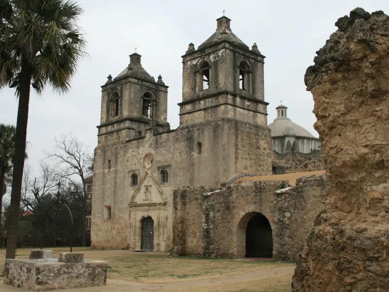 A weathered stone mission church with twin bell towers and a dome, seen past a ruined wall and palm trees.