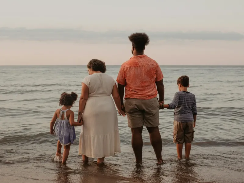 A family of four, seen from behind, holding hands as they wade into a calm sea.