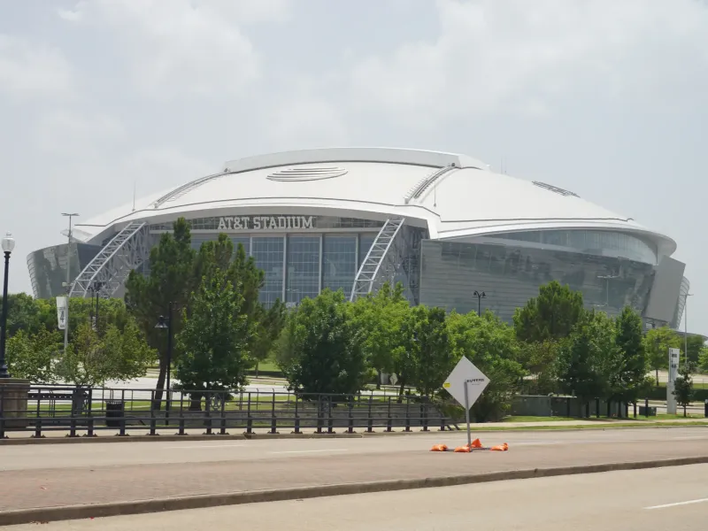 The curved white roof and glass walls of AT&T Stadium in Arlington seen beyond a line of trees.