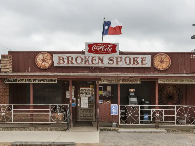 The weathered red front of the Broken Spoke dance hall in Austin with wagon wheels and a Texas flag above the sign.