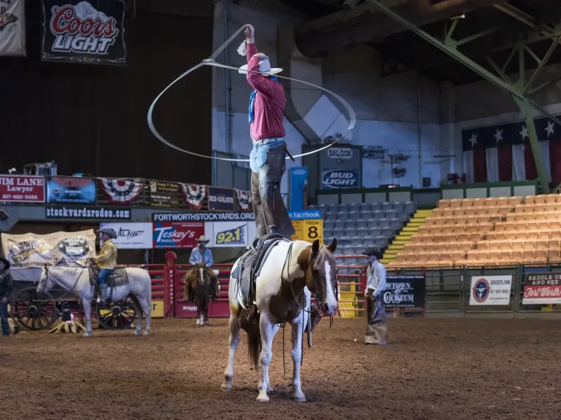 A trick roper stands on a saddled horse and spins a lariat in the dirt arena of the Cowtown Coliseum in Fort Worth.