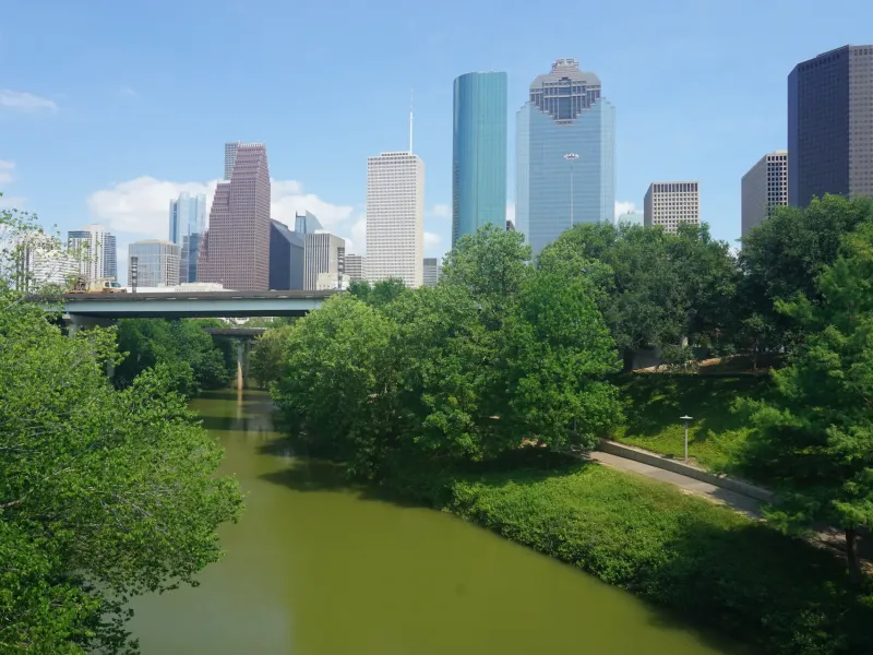Buffalo Bayou winds between wooded banks below the glass towers of the downtown Houston skyline.