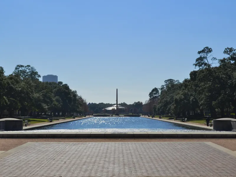 A long reflecting pool framed by live oaks and paved paths stretches toward an obelisk in Hermann Park, Houston.