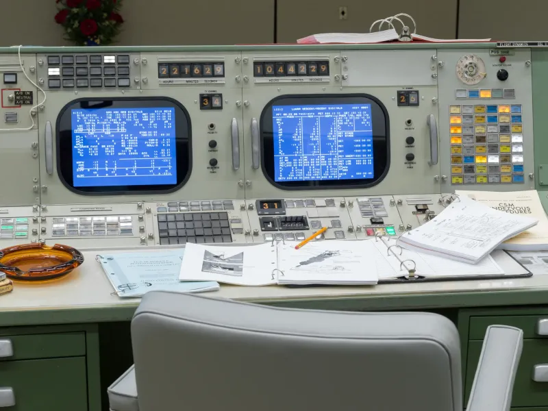 Restored Apollo era consoles with glowing blue screens, switches and open flight binders in Historic Mission Control.