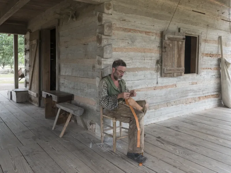 A costumed historical interpreter works a leather strap on the porch of a log farmhouse at George Ranch Historical Park.