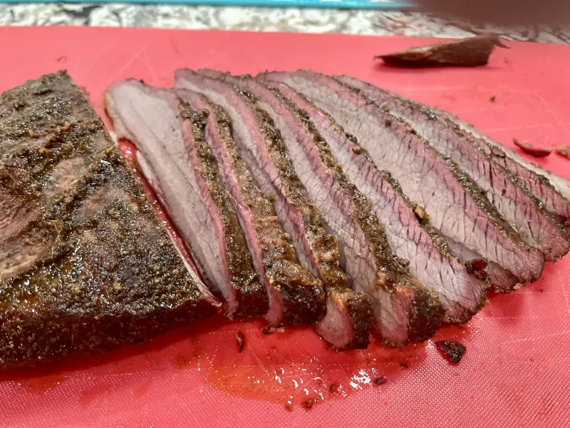 Slices of smoked brisket with a dark peppery bark and a pink smoke ring on a red cutting board.