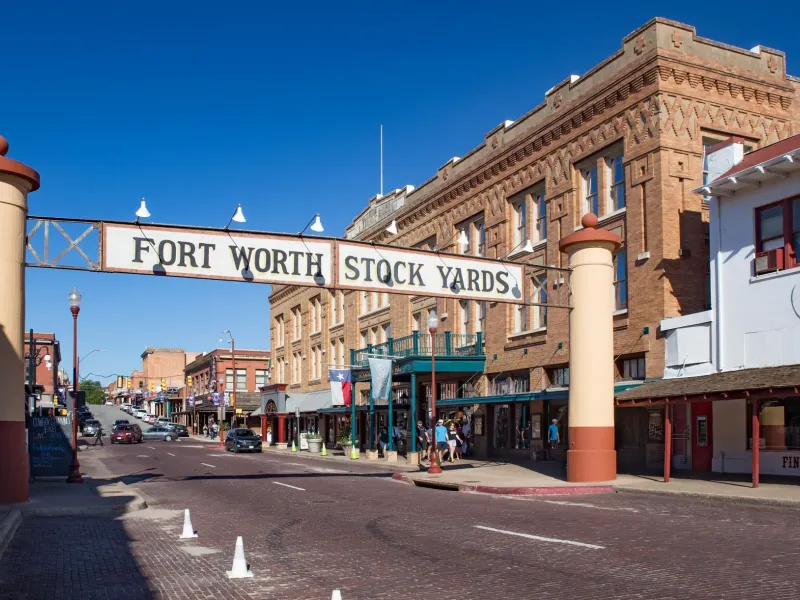 The Fort Worth Stock Yards sign spans a brick street lined with historic buildings under a deep blue sky.
