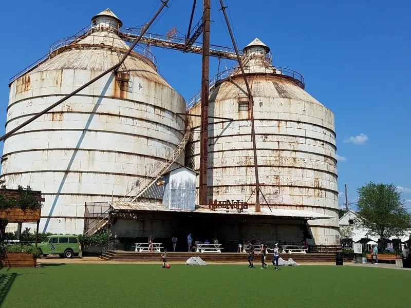 Two weathered steel grain silos rise over a lawn and market stalls at Magnolia Market in Waco, Texas.