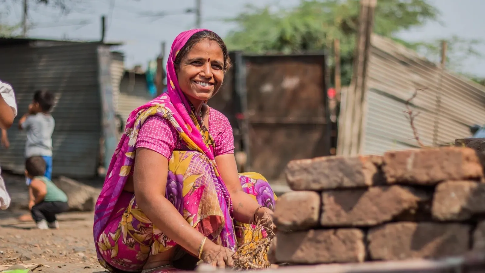 A woman is smiling while sitting on the ground in a rural setting in India featured