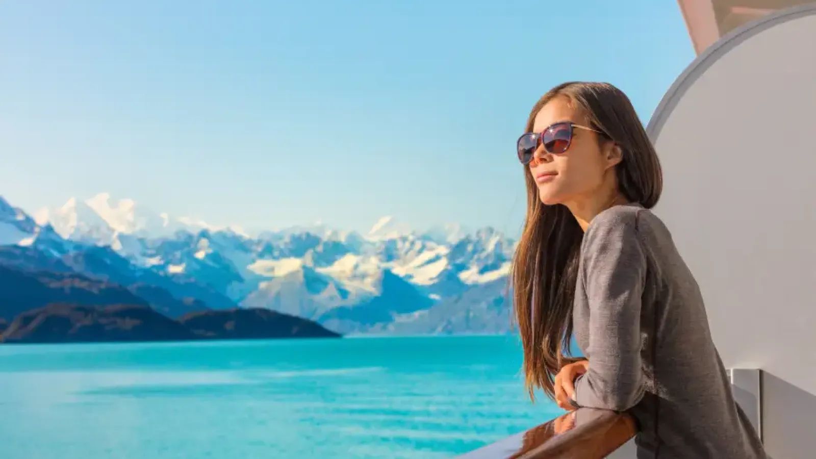 A traveler on a cruise ship balcony in Alaska looking out over snow-capped mountains and glacial water.