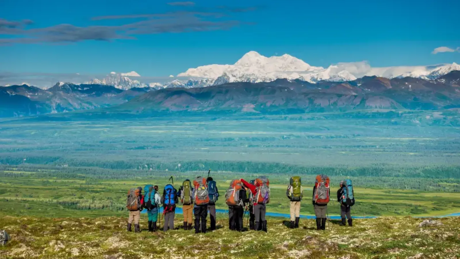 Group of backpackers standing on a tundra ridge overlooking a vast Alaska valley with Denali's snow-capped peak in the bac
