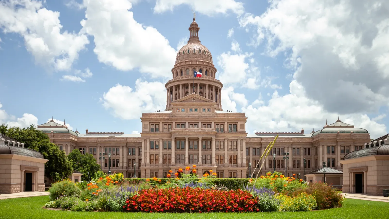 Austin Texas State Capitol building with copper dome Texas flag and colorful flower gardens under a partly cloudy sky