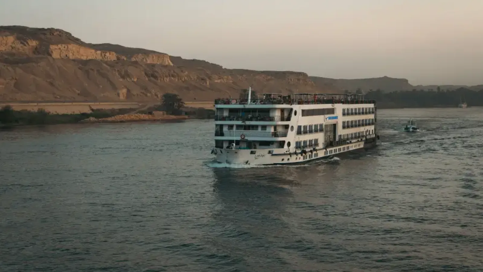 Multi-deck river cruise ship sailing the Nile at dusk with desert cliffs and palm trees in the background.
