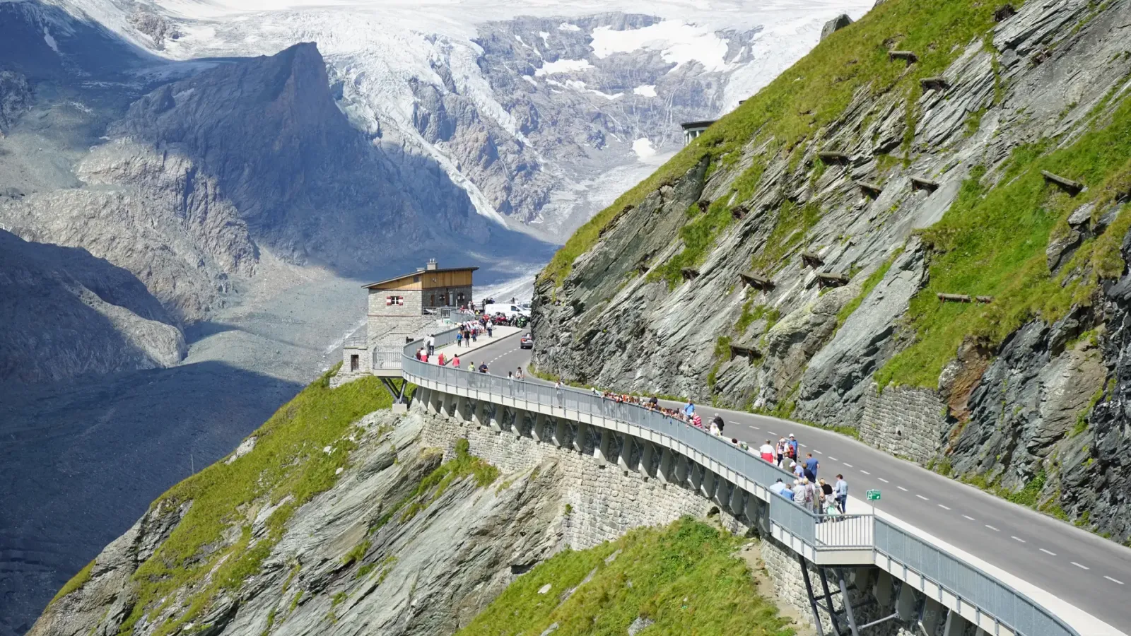 The Grossglockner High Alpine Road curving along a steep green slope with cyclists on it and a glaciated summit behind.