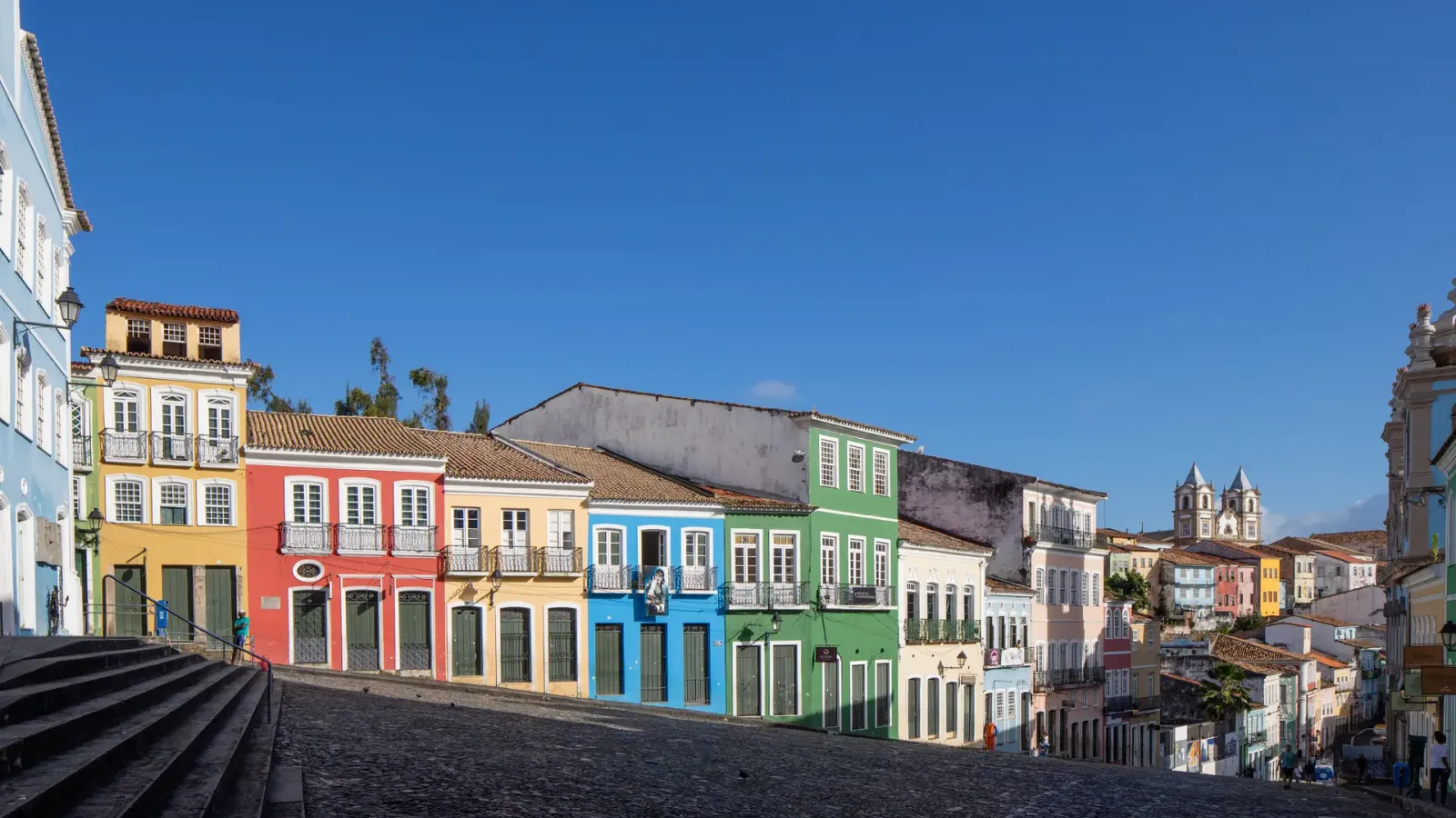 The painted colonial facades of the Largo do Pelourinho in Salvador running down a cobbled slope, with church towers beyond.