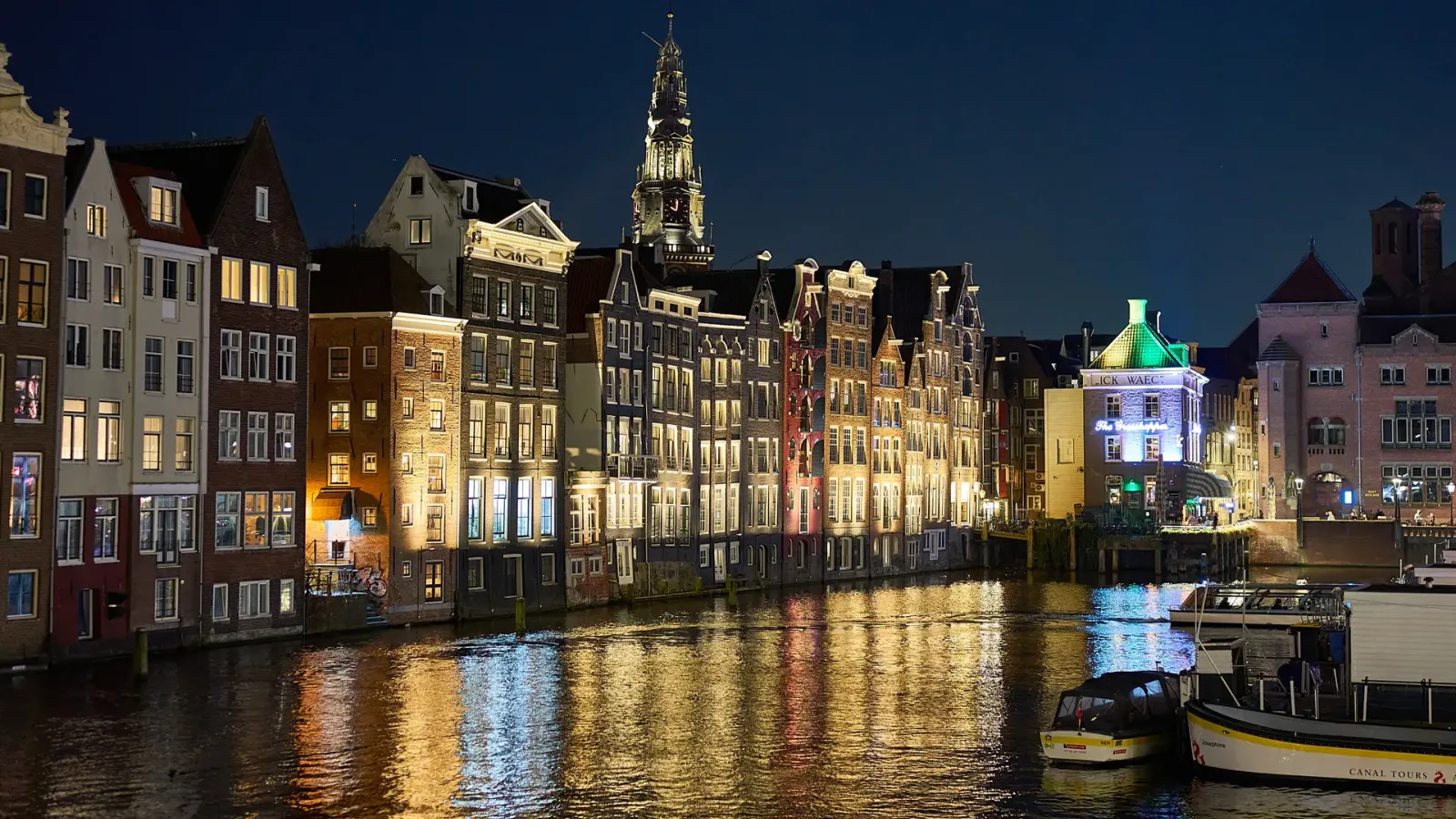 Narrow gabled canal houses reflected in the Damrak in Amsterdam at night, with the tower of the Oude Kerk behind
