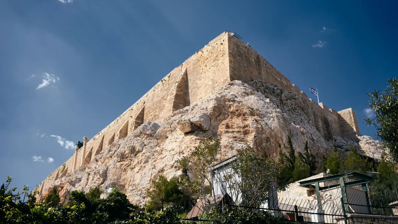 The fortified south wall of the Acropolis of Athens rising above Plaka, with the Greek flag flying at the summit.