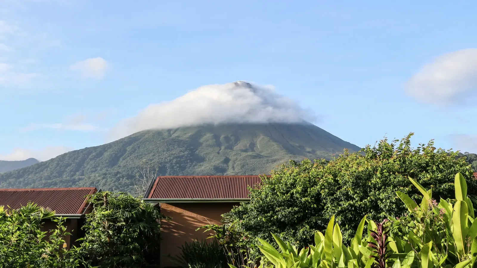 The forested cone of Arenal Volcano in Costa Rica under a cap of cloud, seen above tiled lodge roofs and tropical planting