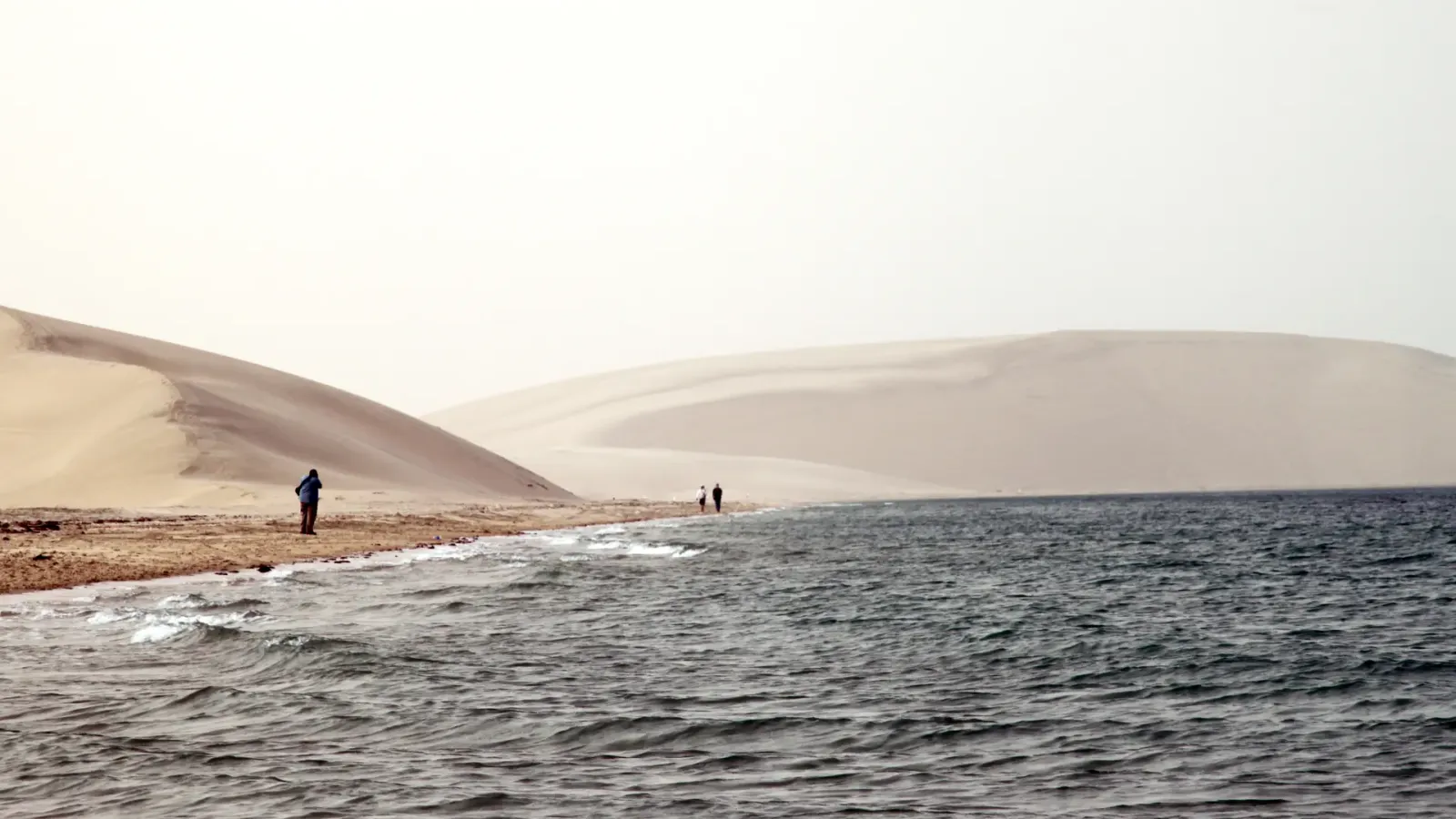 Pale dunes running down to the shallow water of the inland sea at Khor Al Adaid, with two figures at the waterline