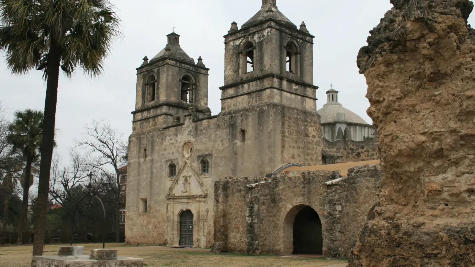 A weathered stone mission church with twin bell towers and a dome, seen past a ruined wall and palm trees.