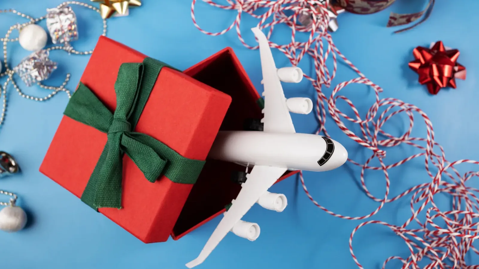 A white toy airplane rising out of an open red gift box tied with green ribbon, among holiday decorations.