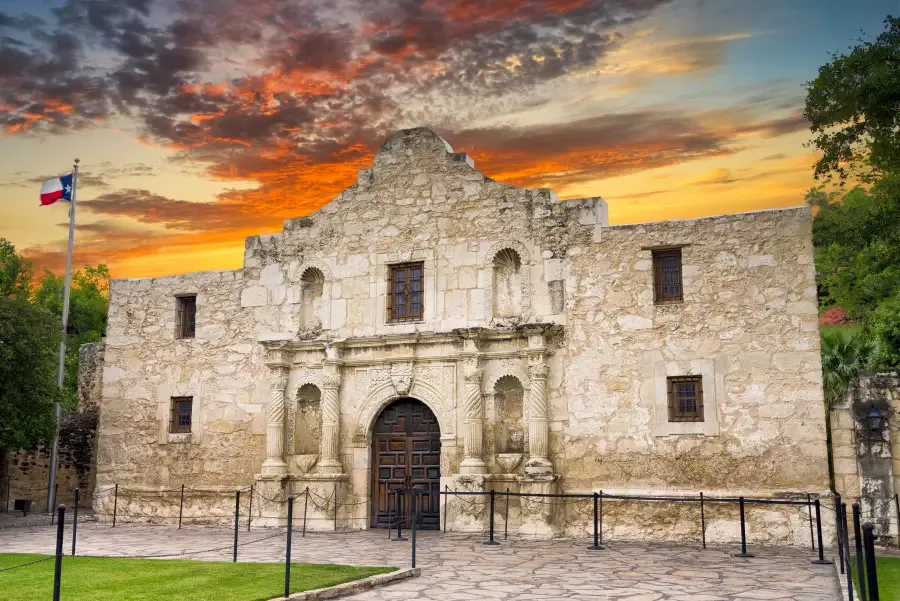 The limestone facade of the Alamo under a sunset sky, with a Texas flag flying beside it.