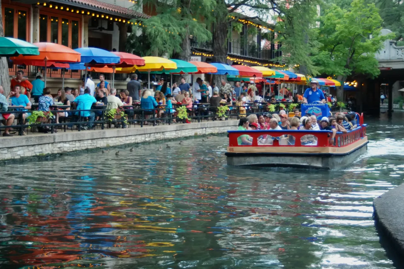A river barge full of seated passengers passing riverside cafe tables under colorful umbrellas on the San Antonio River Walk.