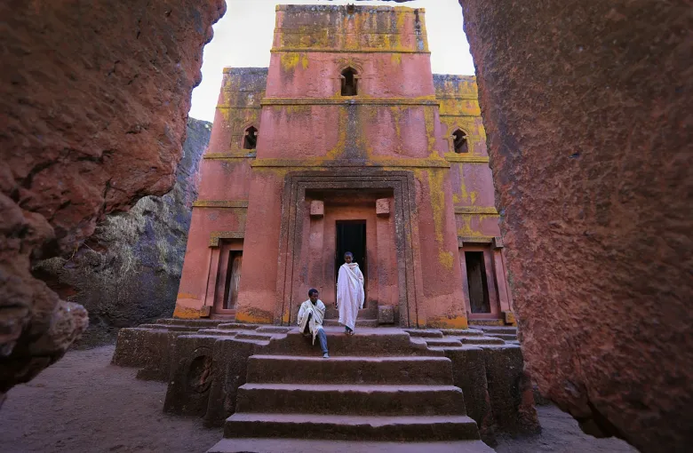 A rock-hewn church at Lalibela, Ethiopia, cut down into reddish rock so that its facade stands at the foot of an open trench, with steps leading down and a figure in white in the doorway.