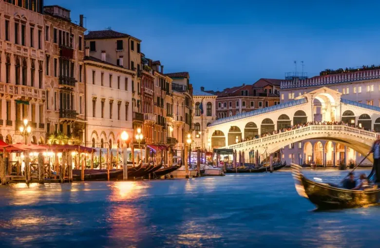 Gondola on the Grand Canal at dusk, with the lit arcades of the Rialto Bridge behind