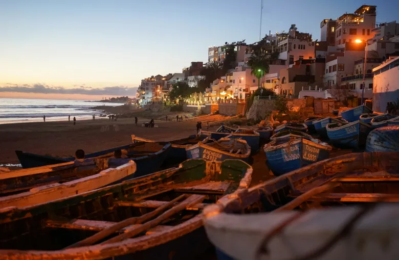 Wooden fishing boats drawn up on the sand at Taghazout, with the town lit at dusk behind.