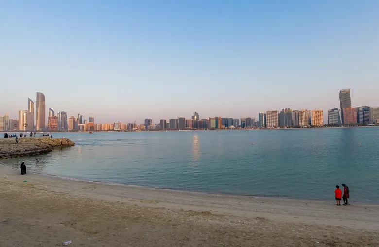 The Abu Dhabi corniche skyline seen across shallow water from a public beach at dusk