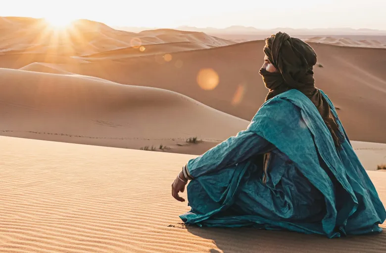 A person in a blue robe and turban sitting on a dune at sunrise in the Erg Chebbi sand sea.
