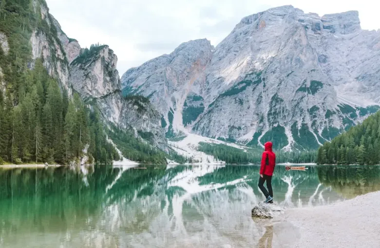 A person in a red jacket beside a still alpine lake below snow-streaked Dolomite peaks