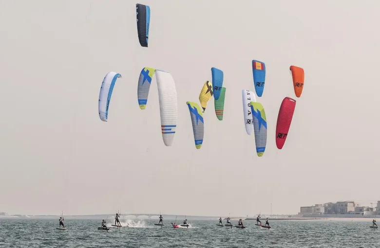 Kitefoil racers on flat coastal water under a hazy sky, with low villas along the shore