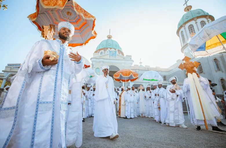Orthodox clergy in white ceremonial robes beneath fringed ceremonial umbrellas, processing in front of a domed church.